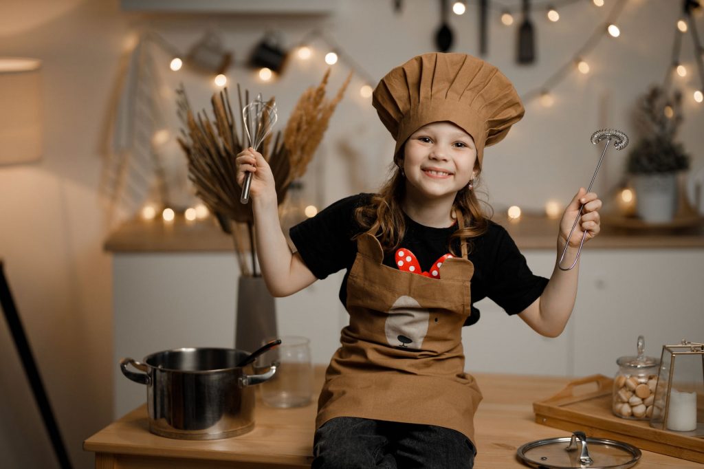 a girl in brown apron and chef hat sitting on a wooden table holding baking equipment while smiling at the camera