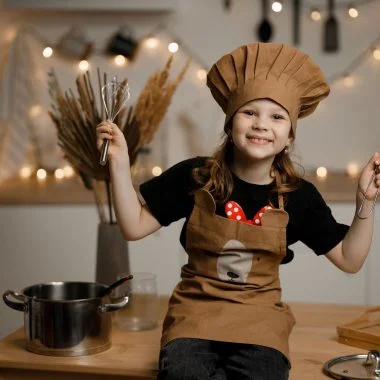 a girl in brown apron and chef hat sitting on a wooden table holding baking equipment while smiling at the camera